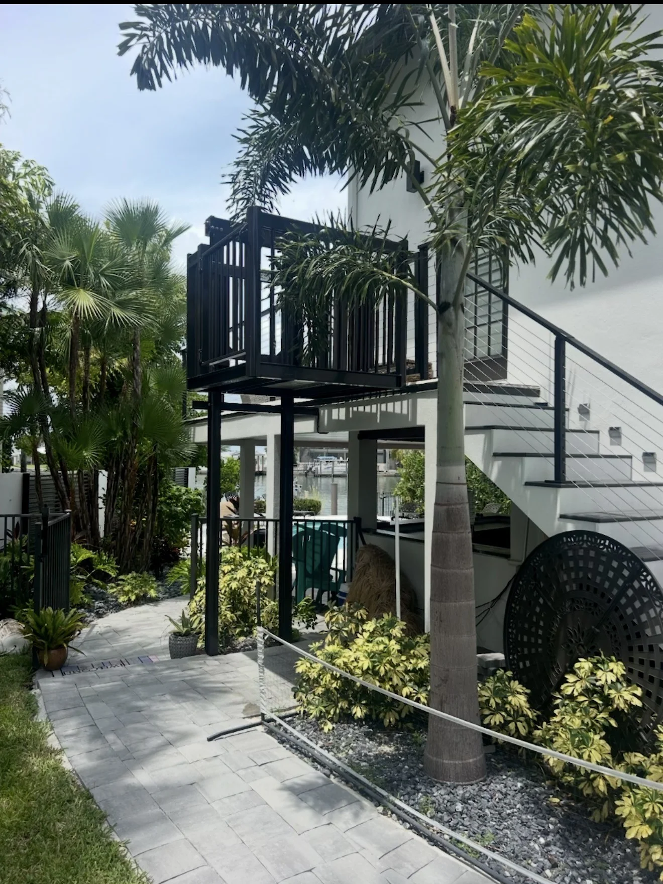 Black steel residential elevator with cable railings on concrete landing beside white house